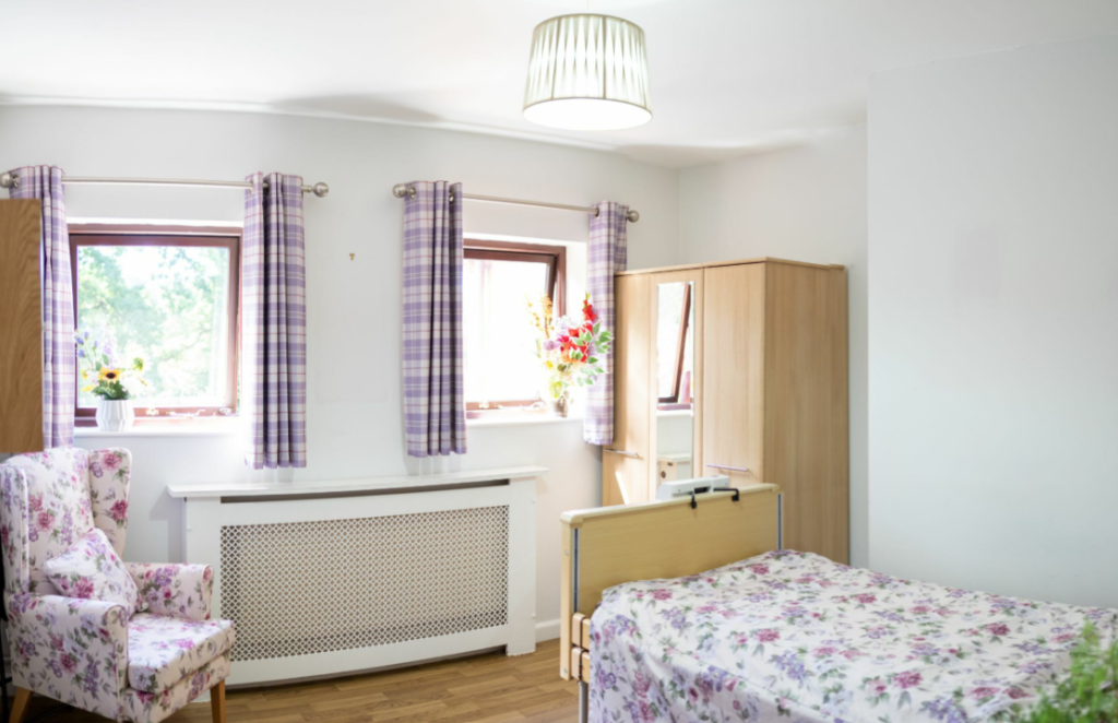 A bright, tidy bedroom with a single bed, floral armchair, wooden wardrobe, and two windows with checked curtains. A lamp hangs from the ceiling.