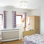 A bright, tidy bedroom with a single bed, floral armchair, wooden wardrobe, and two windows with checked curtains. A lamp hangs from the ceiling.