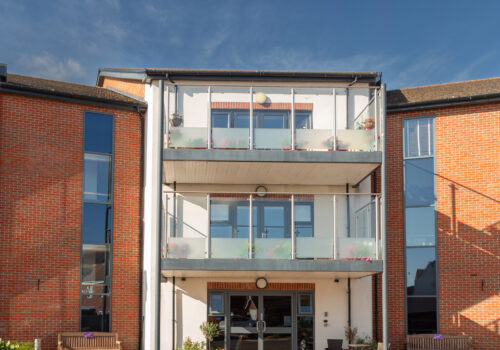 Modern brick building with glass balconies, large windows, and a main entrance welcomes you to Layston Grove, featuring benches outside beneath a clear blue sky.