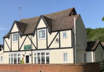 Two-storey cream-coloured building with black timber accents, white windows, and a brown tiled roof, behind a low brick wall, under a clear blue sky.