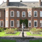 A large brick Georgian-style house with many white-framed windows, a central fountain, and landscaped gardens in the foreground.