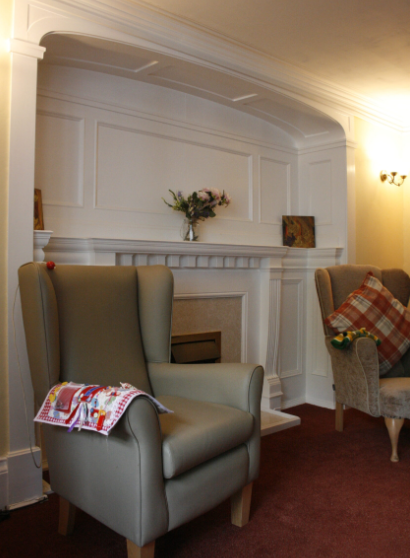 A cosy living room with two armchairs, a fireplace with a flower vase and framed pictures on the mantelpiece, and a check cushion on one chair.