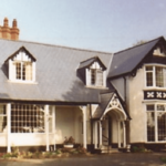 A two-storey house with a slate roof, multiple chimneys, large bay windows, and black-and-white Tudor-style trim surrounded by trees.