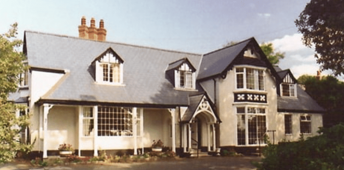A two-storey house with a slate roof, multiple chimneys, large bay windows, and black-and-white Tudor-style trim surrounded by trees.