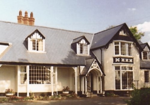 A two-storey house with a slate roof, multiple chimneys, large bay windows, and black-and-white Tudor-style trim surrounded by trees.