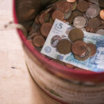 A tin filled with assorted coins, accompanied by a five-pound British banknote, sits on a wooden surface—a simple reminder of days past and perhaps the importance of every penny when Pension Credit comes into play.