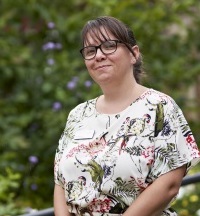 A woman with glasses, wearing a floral shirt, stands outdoors with greenery in the background.