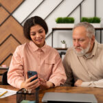 Two people sit at a desk; one shows the other something on a smartphone, perhaps comparing home care costs. A laptop and papers are scattered across the desk.