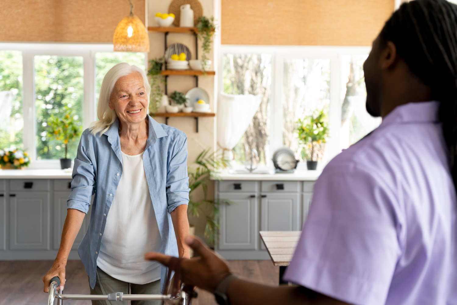 An elderly woman with a walker beams as she chats with a healthcare worker in a bright room, illustrating the warmth of in-home care surrounded by large windows and flourishing plants.