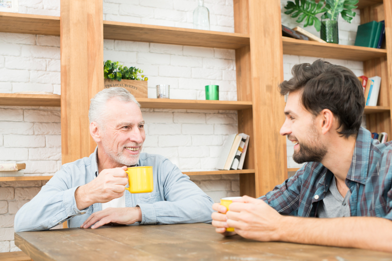 Two men sit at a wooden table, smiling and holding yellow mugs. Surrounding them, bookshelves and plants create a cozy backdrop as they share tips for families dealing with dementia.