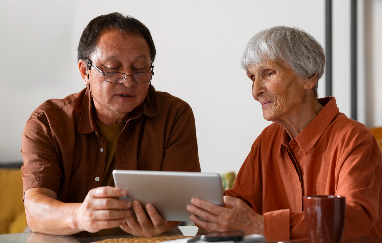 Two people sit at a table, focused on a tablet device displaying information about NHS. Both are wearing glasses and casual clothing, engrossed in understanding continuing healthcare funding.