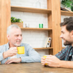 Two men sit at a wooden table, smiling and holding yellow mugs. Surrounding them, bookshelves and plants create a cozy backdrop as they share tips for families dealing with dementia.