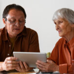 Two people sit at a table, focused on a tablet device displaying information about NHS. Both are wearing glasses and casual clothing, engrossed in understanding continuing healthcare funding.