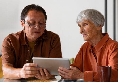 Two people sit at a table, focused on a tablet device displaying information about NHS. Both are wearing glasses and casual clothing, engrossed in understanding continuing healthcare funding.