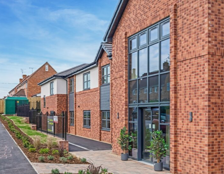 Newly constructed modern brick block of flats with large windows, a paved entrance, and landscaped garden beds near a residential road.