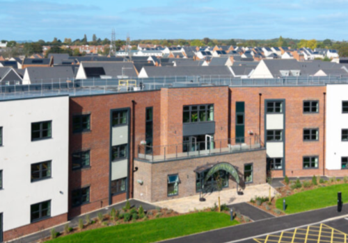 A three-storey modern brick and white building with large windows, an arched entrance, and a car park in front, set in a residential neighbourhood.