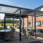 Outdoor patio area with several round tables and wicker chairs under a metal pergola, next to a brick building on a sunny day.