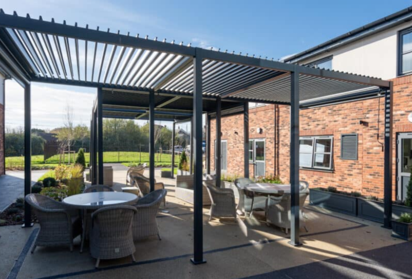 Outdoor patio area with several round tables and wicker chairs under a metal pergola, next to a brick building on a sunny day.
