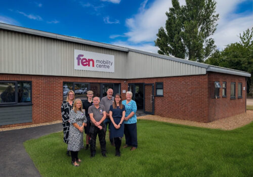 Seven people stand in front of a brick building with a Fen Mobility Centre sign on a sunny day.