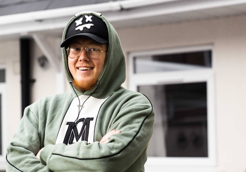 A person wearing glasses, a green hoodie, and a black cap with a cartoon face stands outside with arms folded, smiling at the camera.