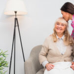 A smiling elderly woman sits in an armchair in a care home whilst a younger woman stands beside her, kissing her on the forehead and holding her hand. A standard lamp and plant are in the background.