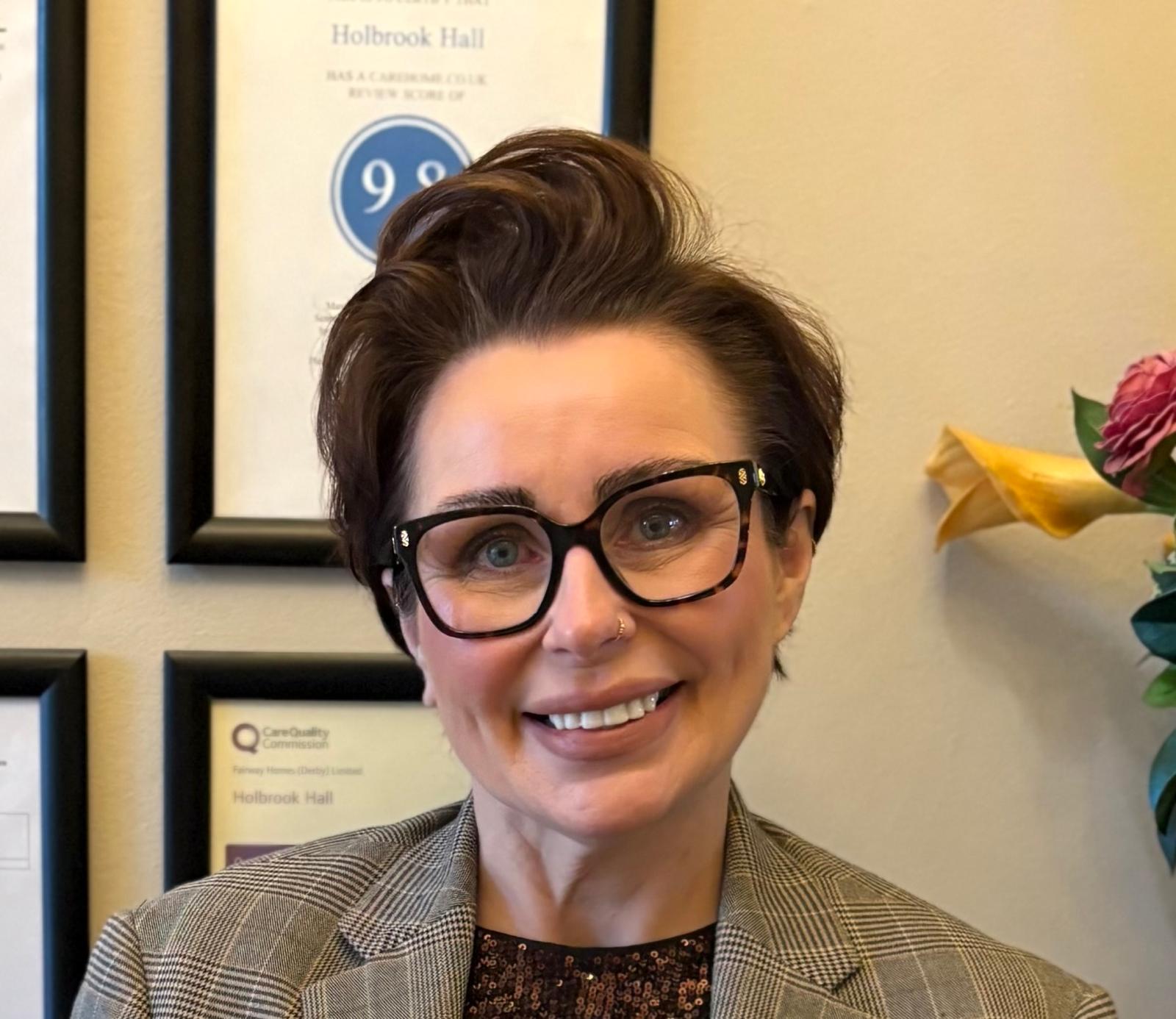A person with short brown hair, glasses, and a nose ring smiles at the camera, standing in front of framed certificates and a yellow flower.