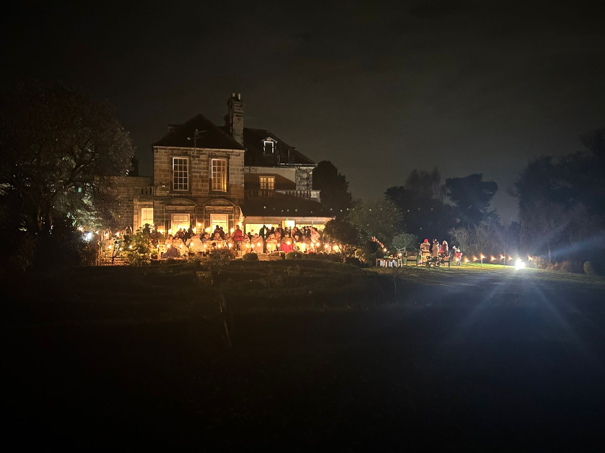 A large house is illuminated at night, with a crowd of people gathered outside under fairy lights; the surrounding garden is dark.