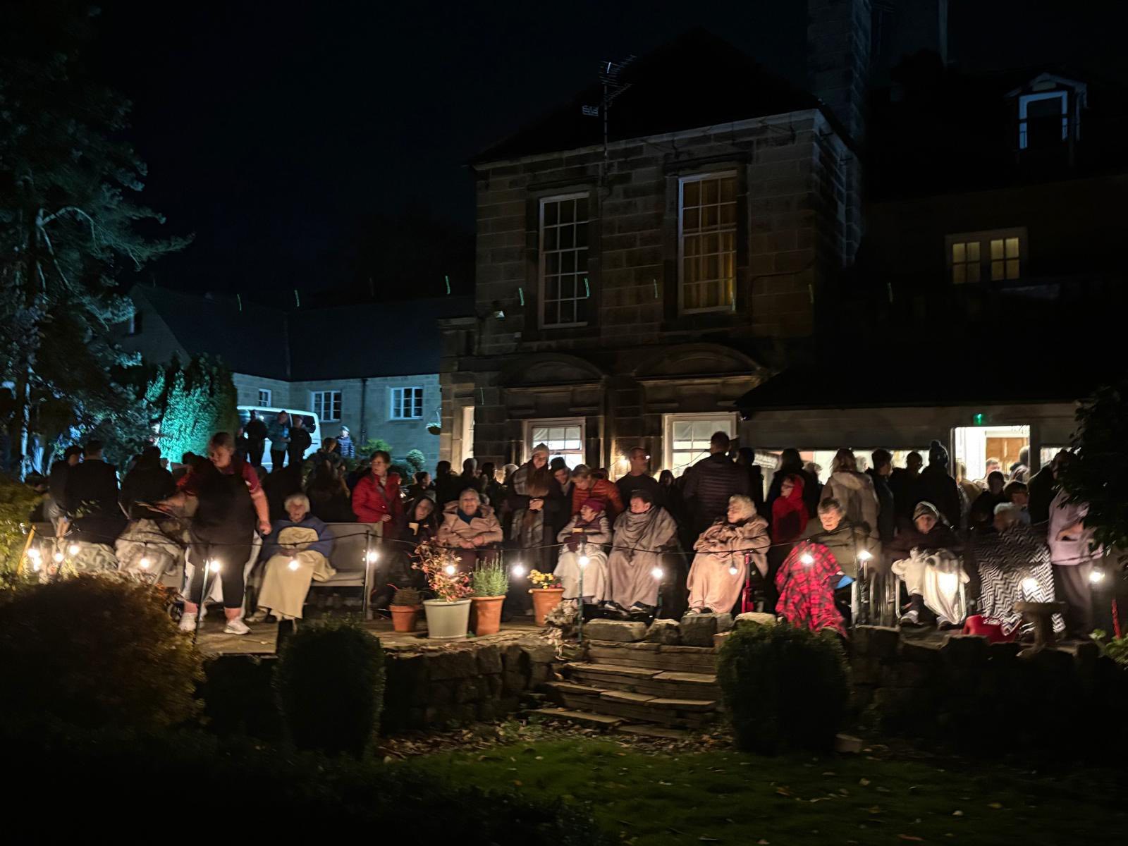 A large group of people, many holding candles, gather outside a building at night. Potted plants and shrubs are visible in the foreground.
