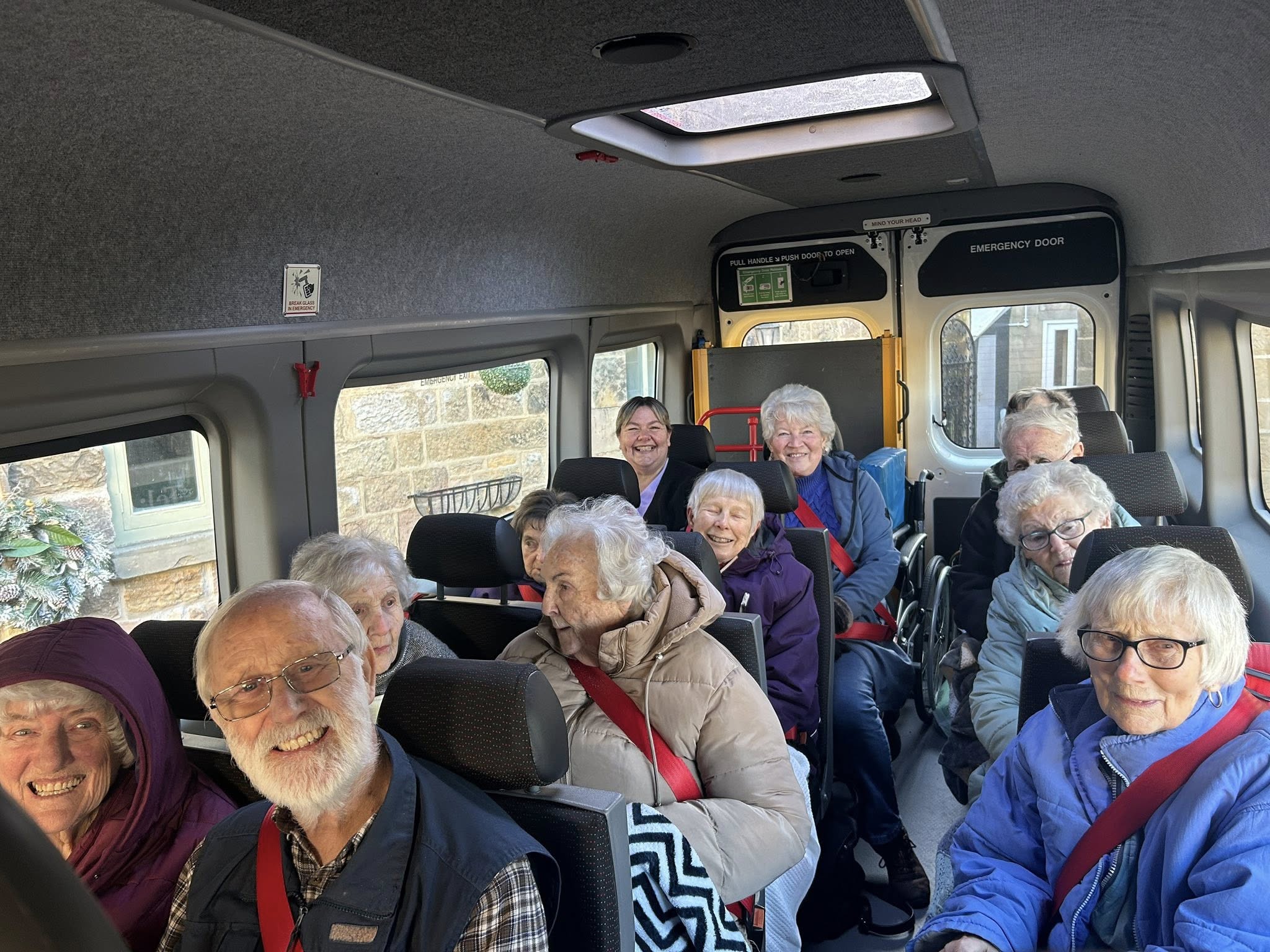 A group of elderly people, all wearing seat belts, sit together and smile inside a minibus.