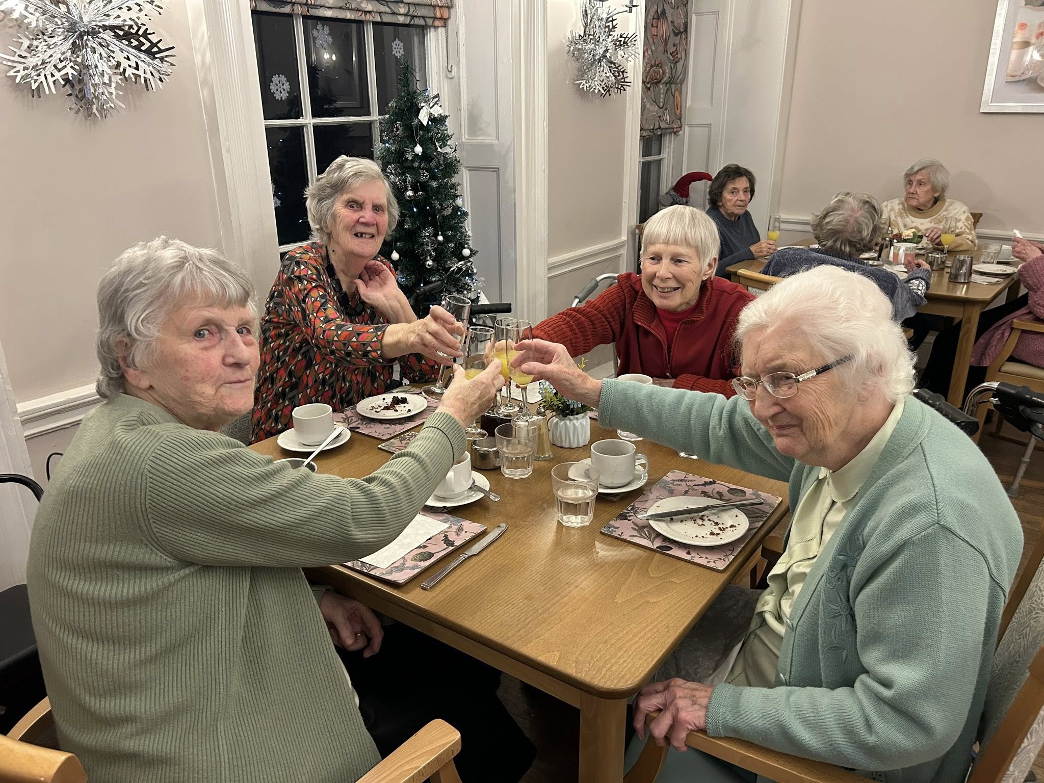 Four elderly women sit at a dining table raising glasses in a toast, with finished plates in front of them and holiday decorations in the background.