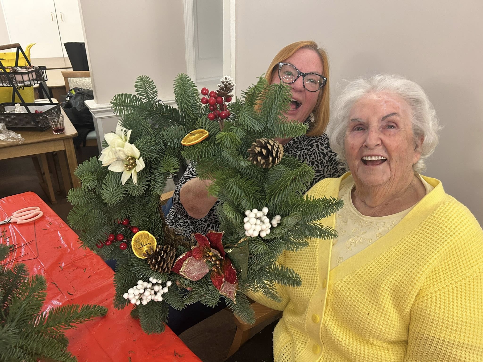 Two women sit at a table holding a decorated holiday wreath made of evergreens, berries, pine cones, and dried citrus slices. Both are smiling.