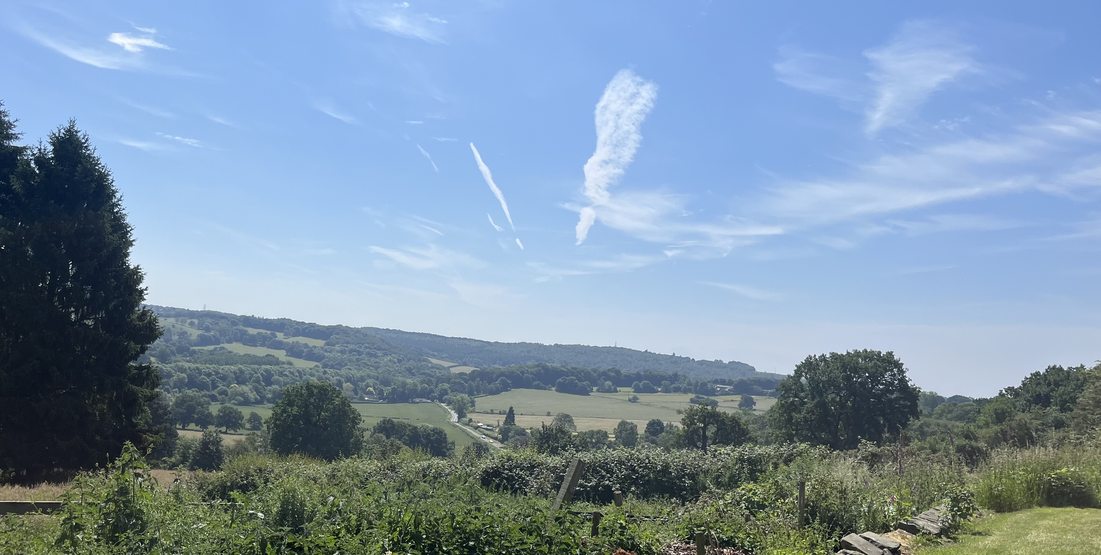 A rural landscape with green fields, trees, and distant hills under a blue sky with scattered white clouds.