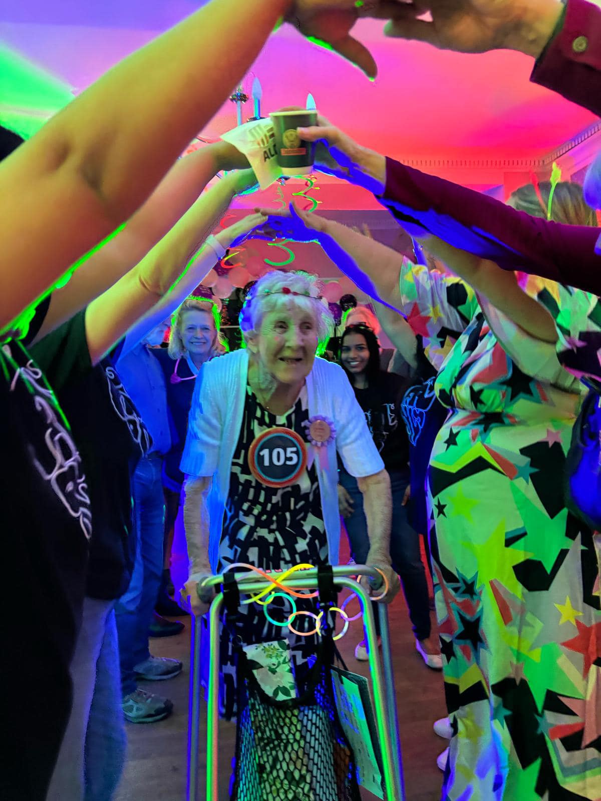 An elderly woman with a walking frame and a 105 badge walks under an arch of raised arms at a colourful indoor celebration.