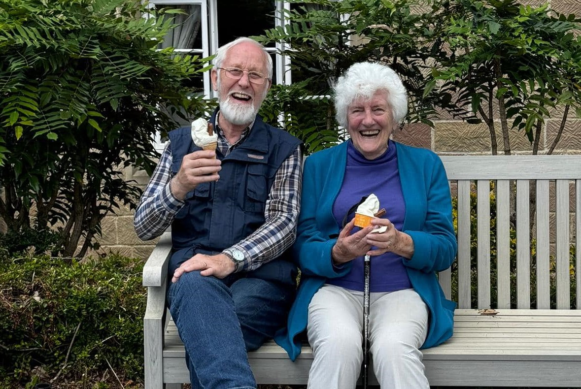 An older man and woman sit on a bench outdoors, smiling and holding ice cream cones. Shrubs and a stone building are visible in the background.