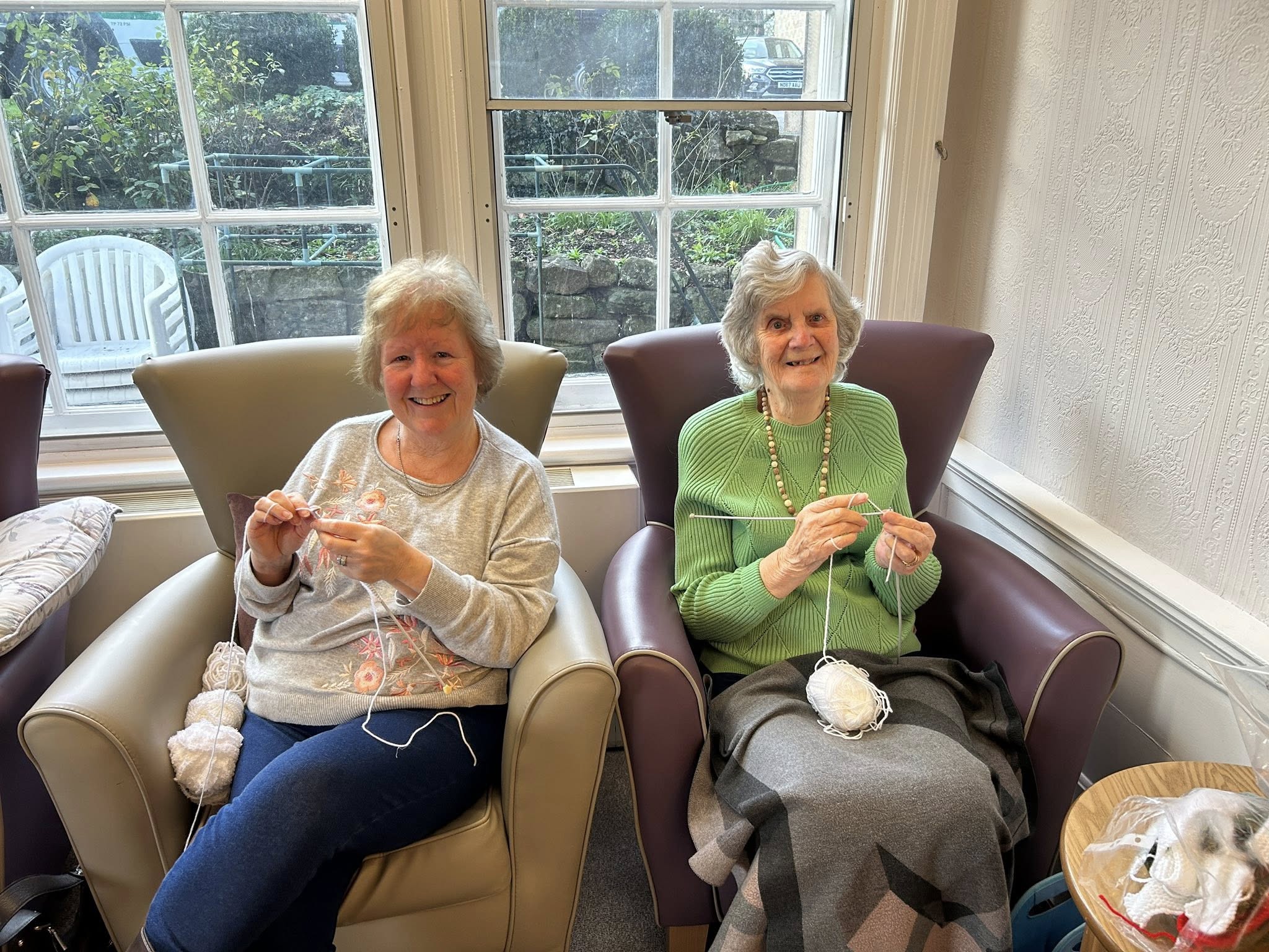 Two elderly women sitting in armchairs by a window, smiling whilst knitting with white wool.