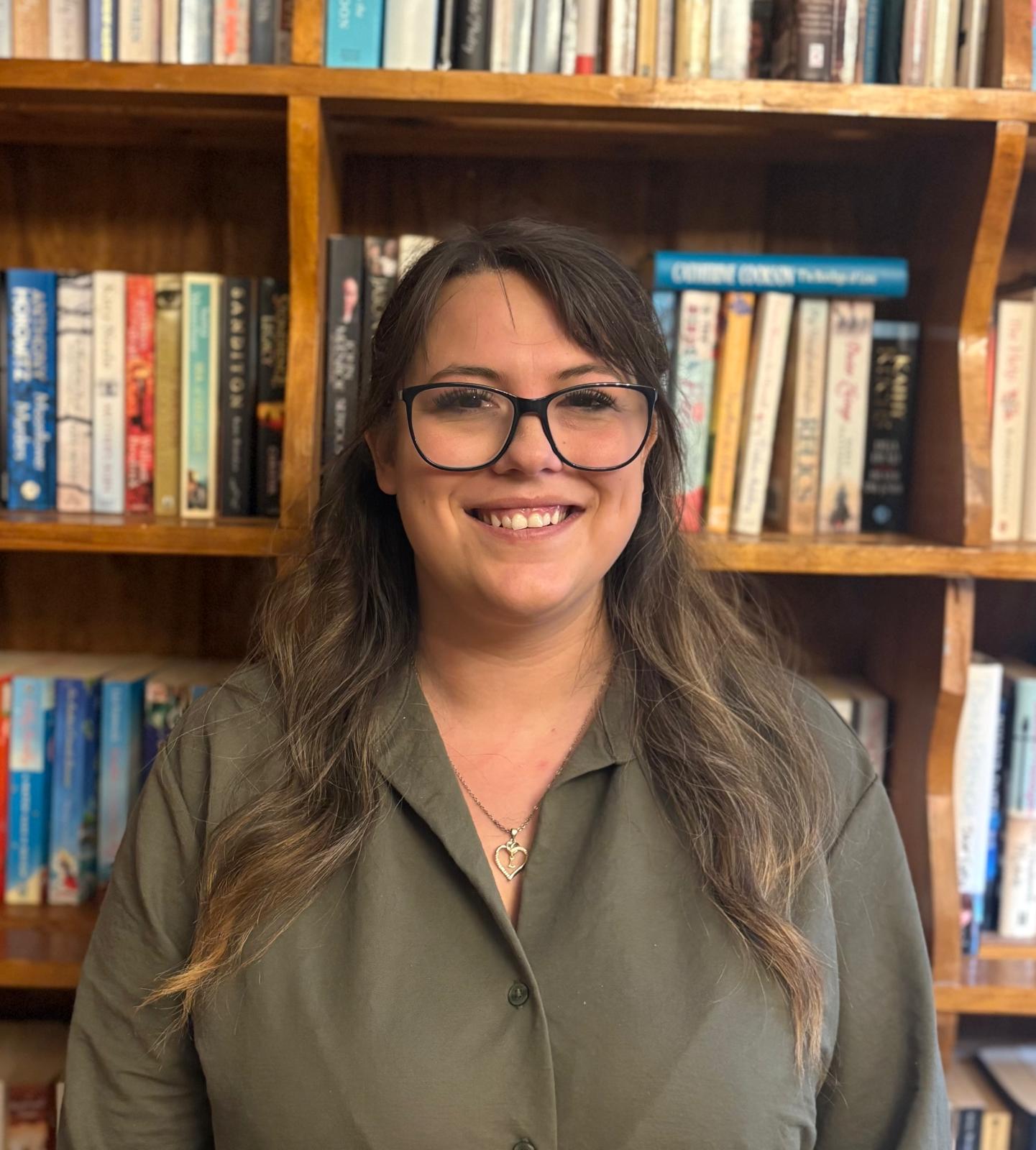 A woman with long brown hair and glasses stands in front of bookshelves filled with books, smiling at the camera. She is wearing a green shirt and a heart-shaped necklace.