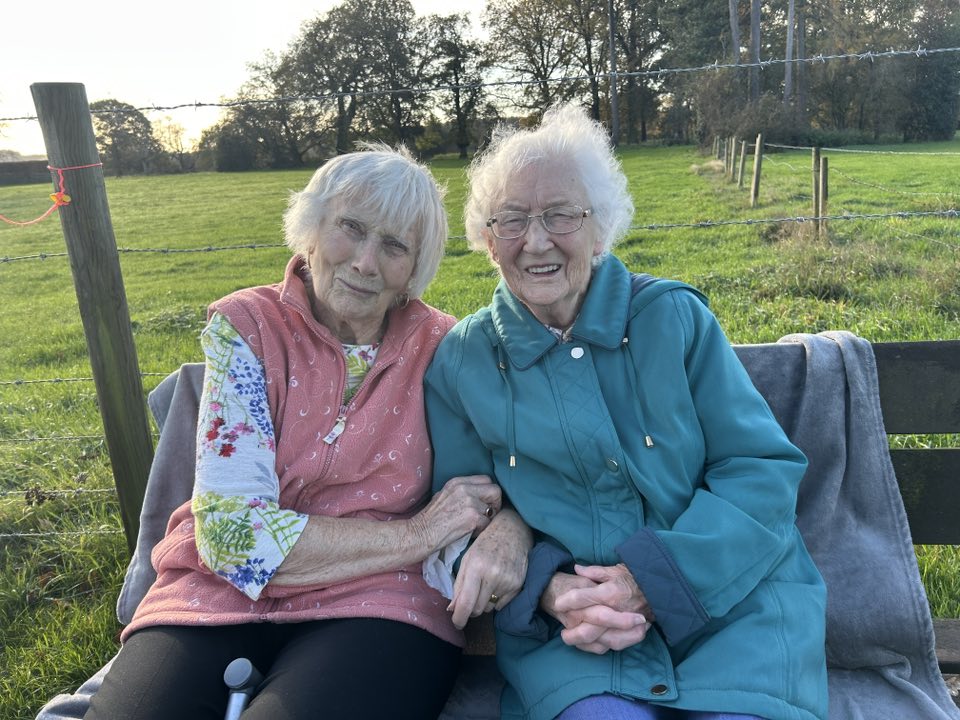 Two elderly women sit closely together on a bench outdoors, smiling at the camera, with a grassy field and trees in the background.