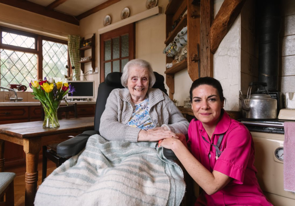 An elderly woman sits in a wheelchair with a blanket on her lap, smiling beside a carer in a pink uniform inside a cosy kitchen.