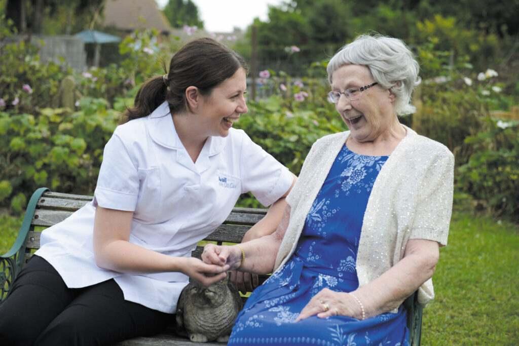 A carer and an elderly woman sit on a bench in a garden, smiling and holding hands, with a grey cat on the woman's lap.