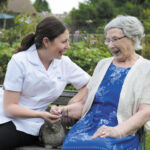 A carer and an elderly woman sit on a bench in a garden, smiling and holding hands, with a grey cat on the woman's lap.