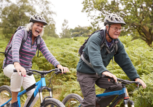 Two older adults wearing helmets and rucksacks ride mountain bikes on a trail through green ferns, with trees in the background.