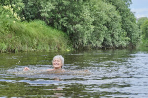 An older woman with short grey hair swims and smiles in a river surrounded by green trees and vegetation.