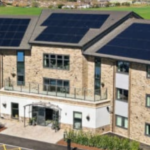 A modern, multi-storey building with solar panels covering most of the roof, surrounded by greenery and a car park in front.
