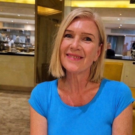 A woman with blonde hair wearing a blue shirt sits and smiles in a restaurant with an open kitchen in the background at Richmond Villages Willaston.