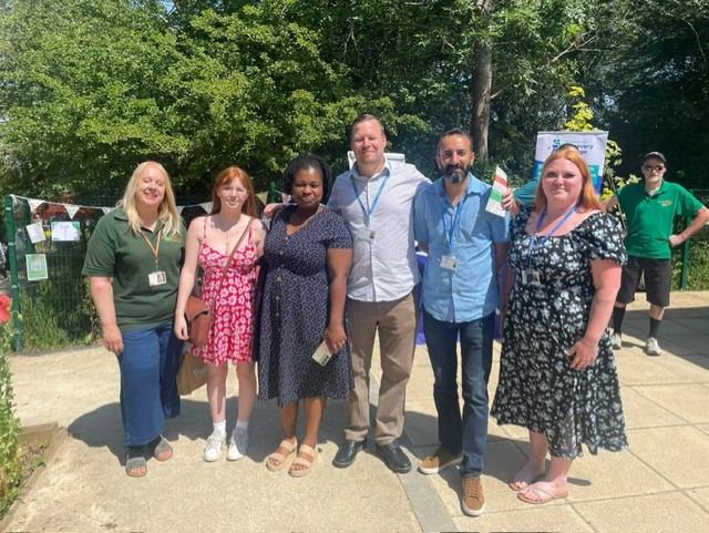 Six adults stand outdoors in sunny weather, posing for a group photo on a paved path with greenery and event signs in the background.