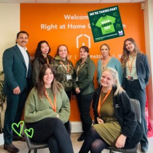 Eight people pose for a group photo in front of an orange wall with Right at Home signage, several wearing green clothing and orange lanyards, with a Wear Green Day sign above.
