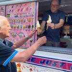 An ice cream vendor hands two soft serve cones to an elderly customer at an ice cream van with a colourful menu on display.