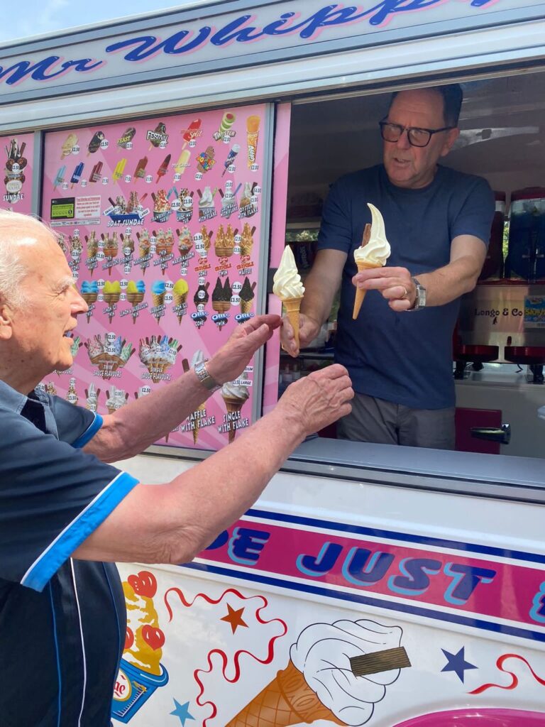 An ice cream vendor hands two soft serve cones to an elderly customer at an ice cream van with a colourful menu on display.