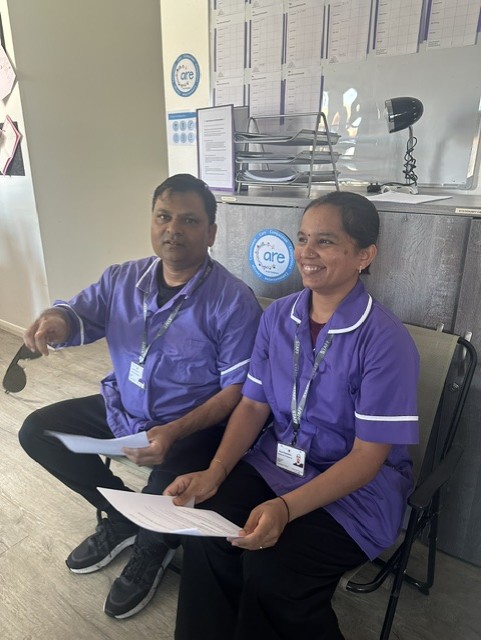 Two healthcare workers in purple uniforms sit indoors, holding papers and smiling. A desk with documents, a lamp, and an award plaque is visible in the background.