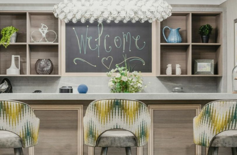 Modern kitchen with a chalkboard that says “Welcome”, decorative shelves, a vase of flowers on the worktop, and three patterned bar stools.